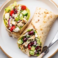 Freshly grilled Greek Lemon Chicken Salad Wrap sliced open on a plate, showing chopped cucumbers, tomatoes, and crumbled feta inside a warm tortilla.